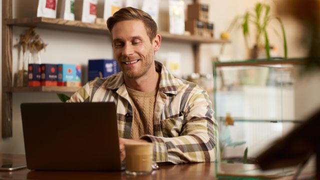 Homem de pele clara e camisa xadrez usando um notebook em mesa de madeira; ao fundo, prateleiras com pacotes e caixas de produtos.