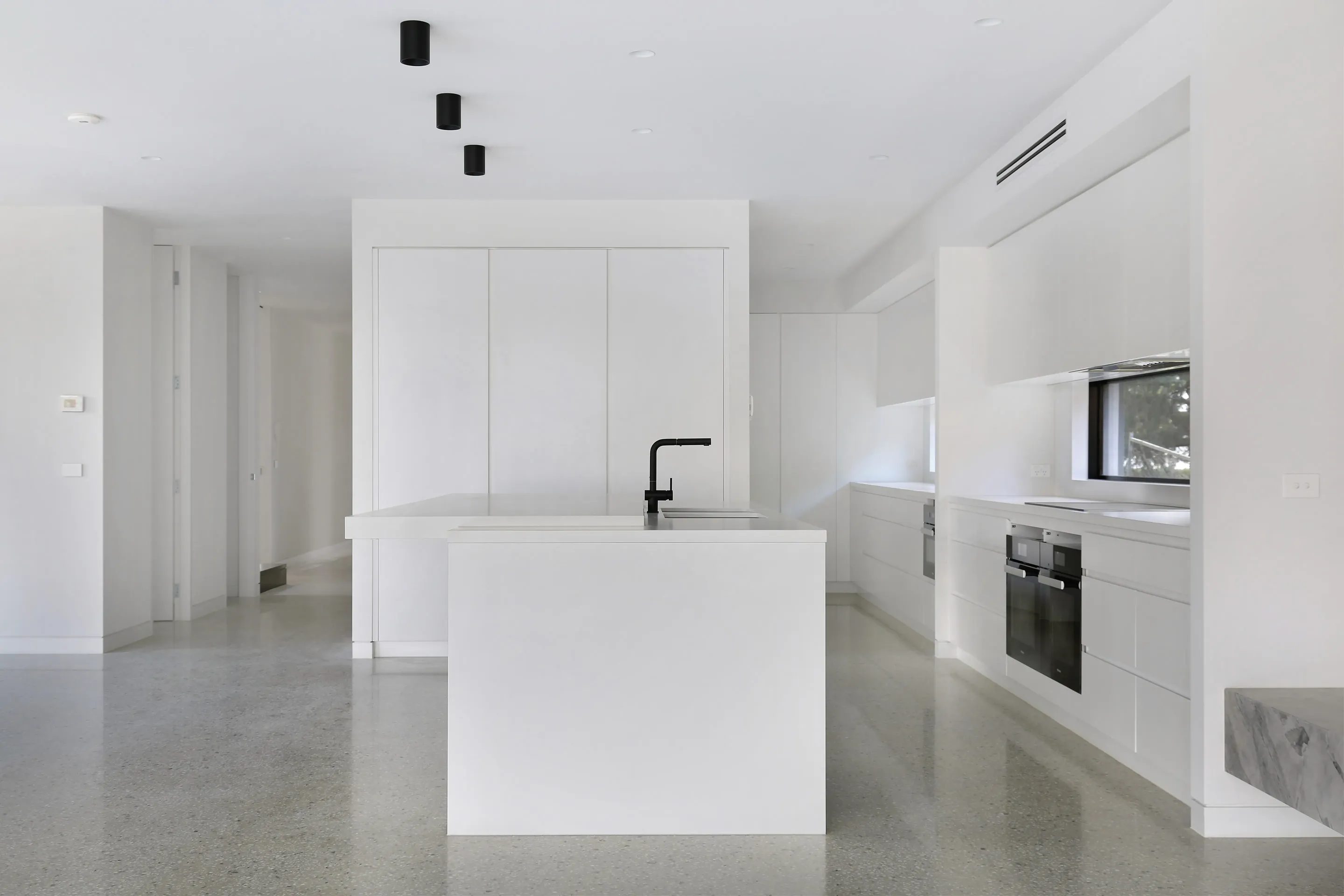 Minimalist white kitchen with a waterfall island and black tap, black surface-mounted downlights, integrated black ovens, an induction cooktop under a window and polished-concrete floors