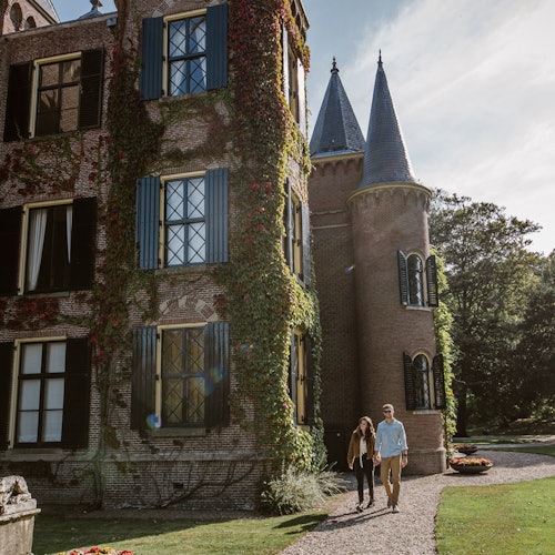 两个人沿着一条碎石小路行走，小路旁是一座历史悠久的砖砌建筑，墙上覆盖着常春藤，还有塔楼。