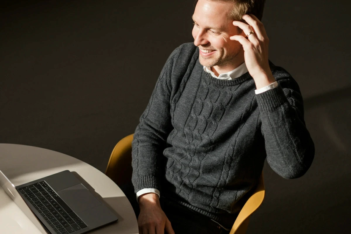 A man in a black sweater smiles while sitting at a table, with a laptop in front of him.