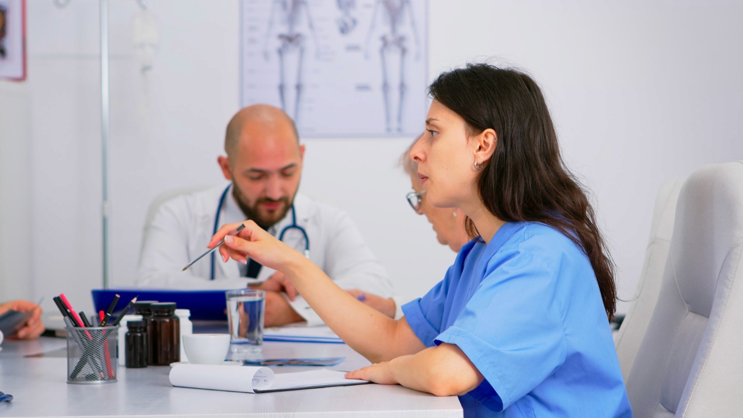A female doctor in blue scrubs gestures while discussing with a male colleague, who has a stethoscope. They are in a bright medical office setting.