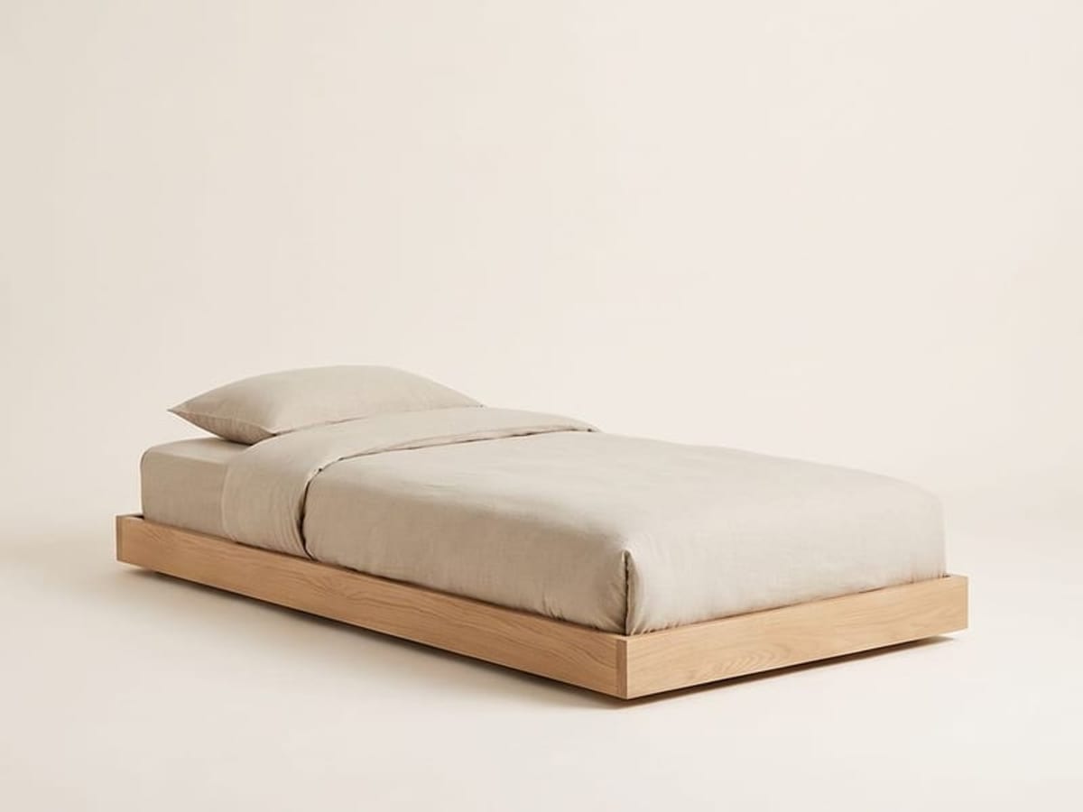Niedriges Einzelbett aus hellem Holz ohne Kopfteil mit sandfarbener Bettwäsche vor hellem Hintergrund