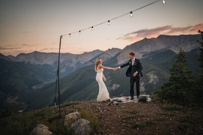 The newlyweds dance beneath string lights with a mountain panorama behind them.
