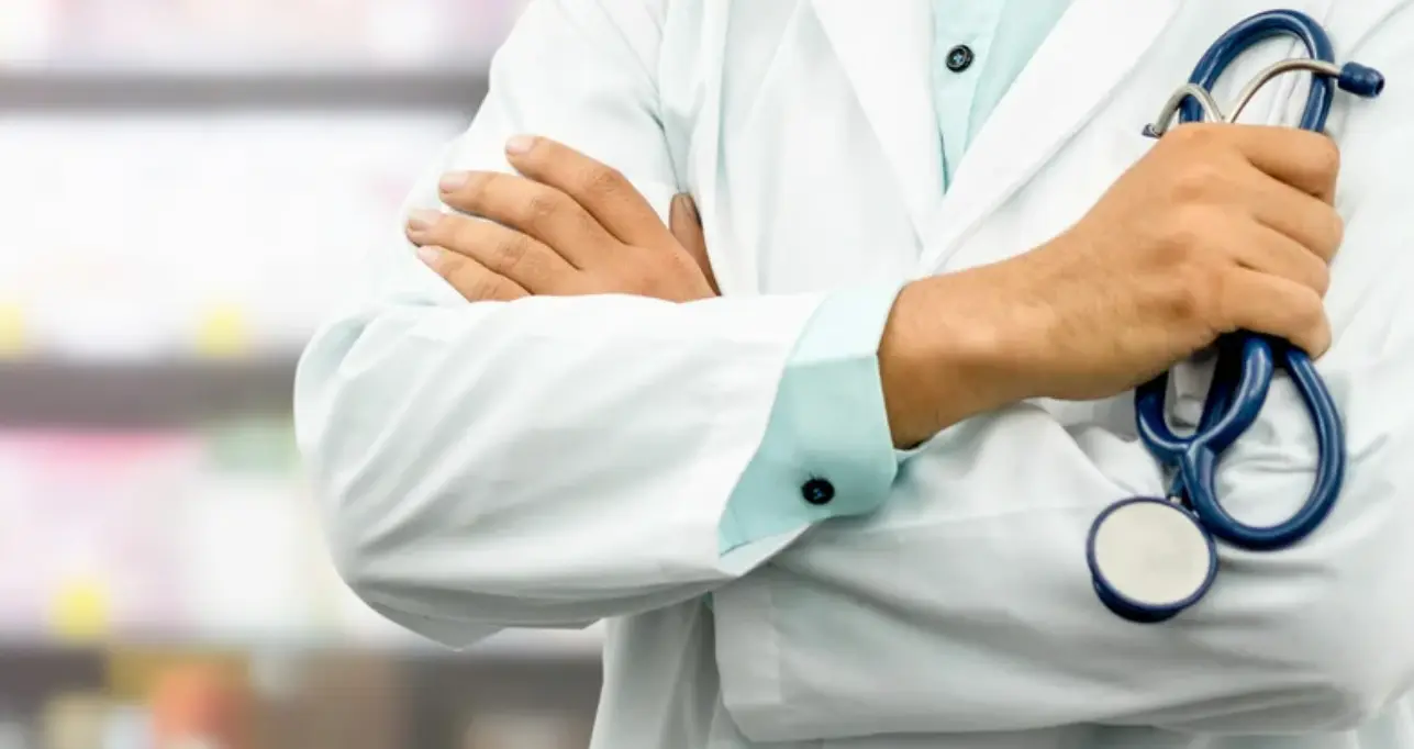 Primo piano di un professionista sanitario in camice bianco con le braccia incrociate che tiene uno stetoscopio.