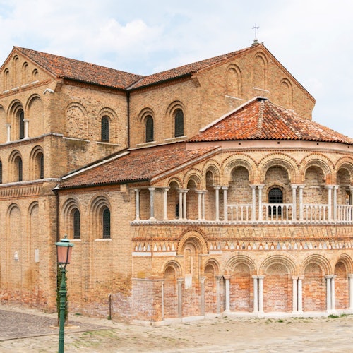 Historic brick building with arched windows, decorative facades, and a tiled roof, set against a partly cloudy sky.