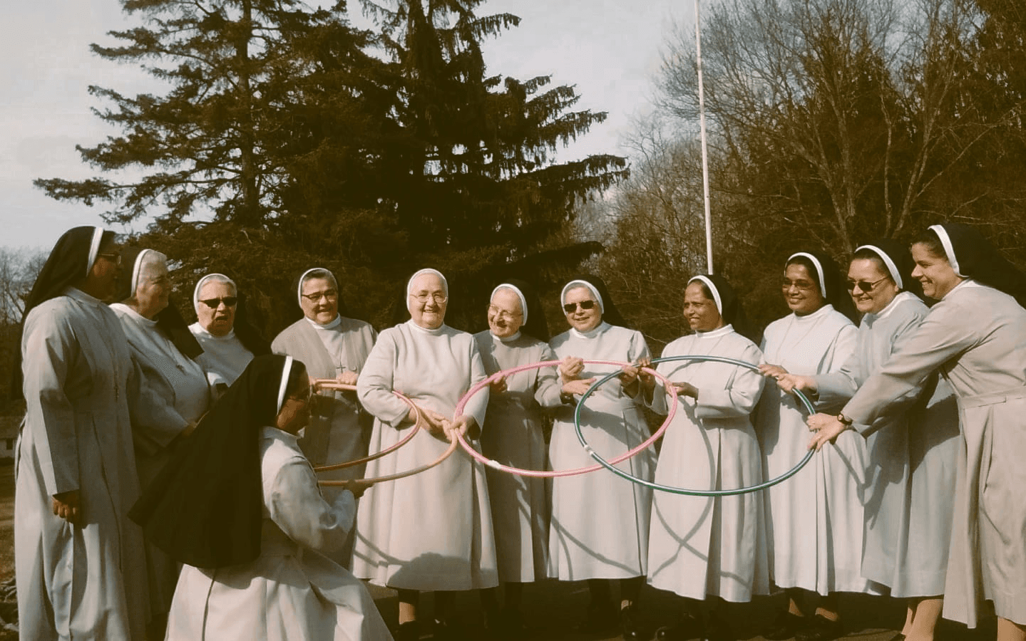 Group of twelve Catholic nuns from the Daughters of Our Lady of the Garden, USA province, standing outdoors in a semicircle. Dressed in light gray habits with black veils, the sisters hold colorful hula hoops in a joyful, recreational moment. Set against a winter landscape with leafless trees and an evergreen, this image reflects the congregation’s charism of Vigilant Evangelical Charity and their commitment to joyful service and community life.