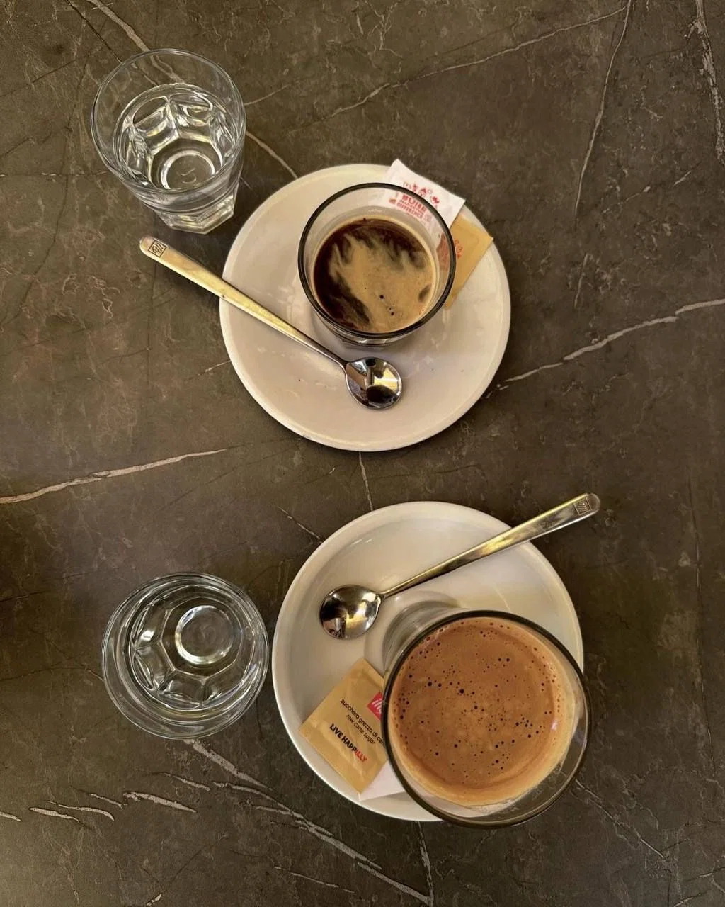 Dos tazas de café expreso con agua y cucharas sobre una mesa de mármol.