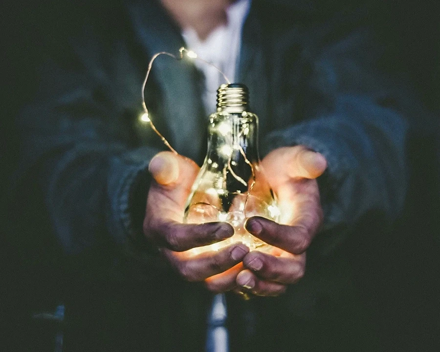 Person holding a lit light bulb filled with string lights in cupped hands.
