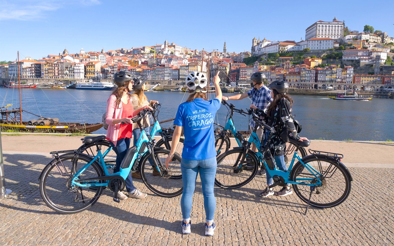 Passeio em grupo de bicicletas elétricas com guia pelo Rio Douro, com a paisagem urbana do Porto ao fundo.