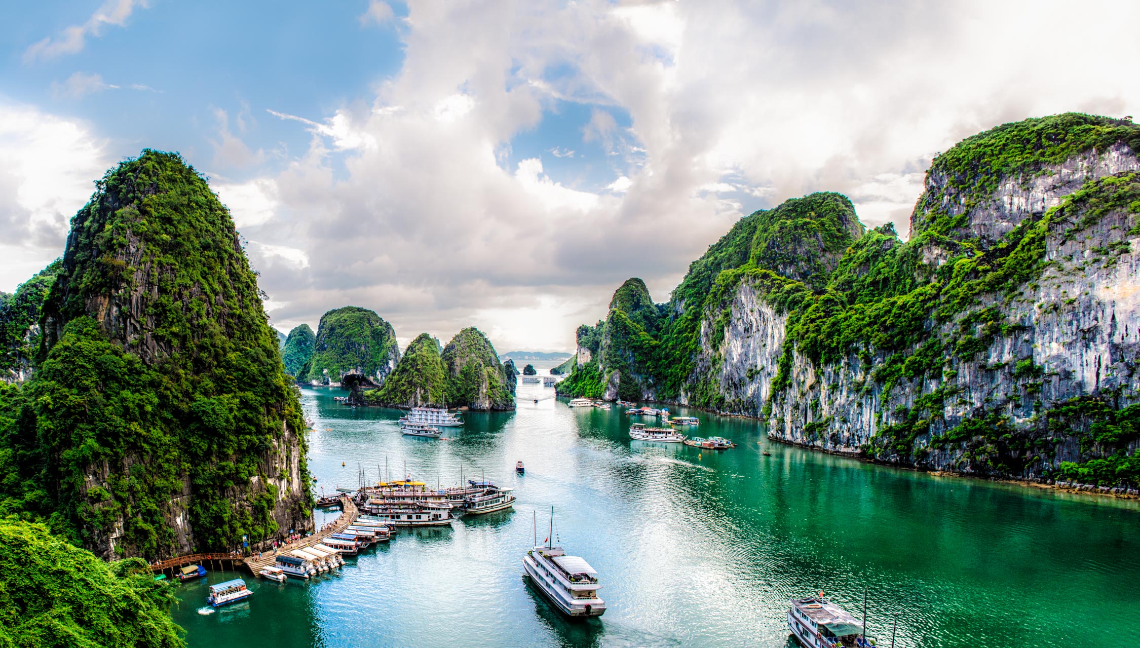 Limestone karsts covered in green vegetation rise from turquoise waters with several boats and a small village nearby.