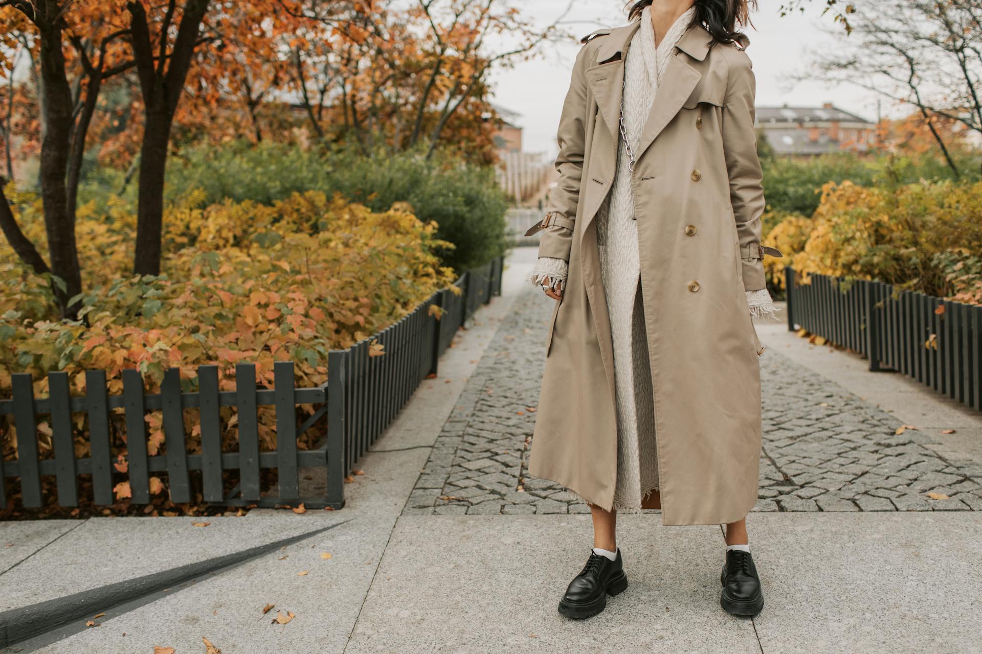 A woman in a trench coat stands on a path and looks up at the autumn trees
