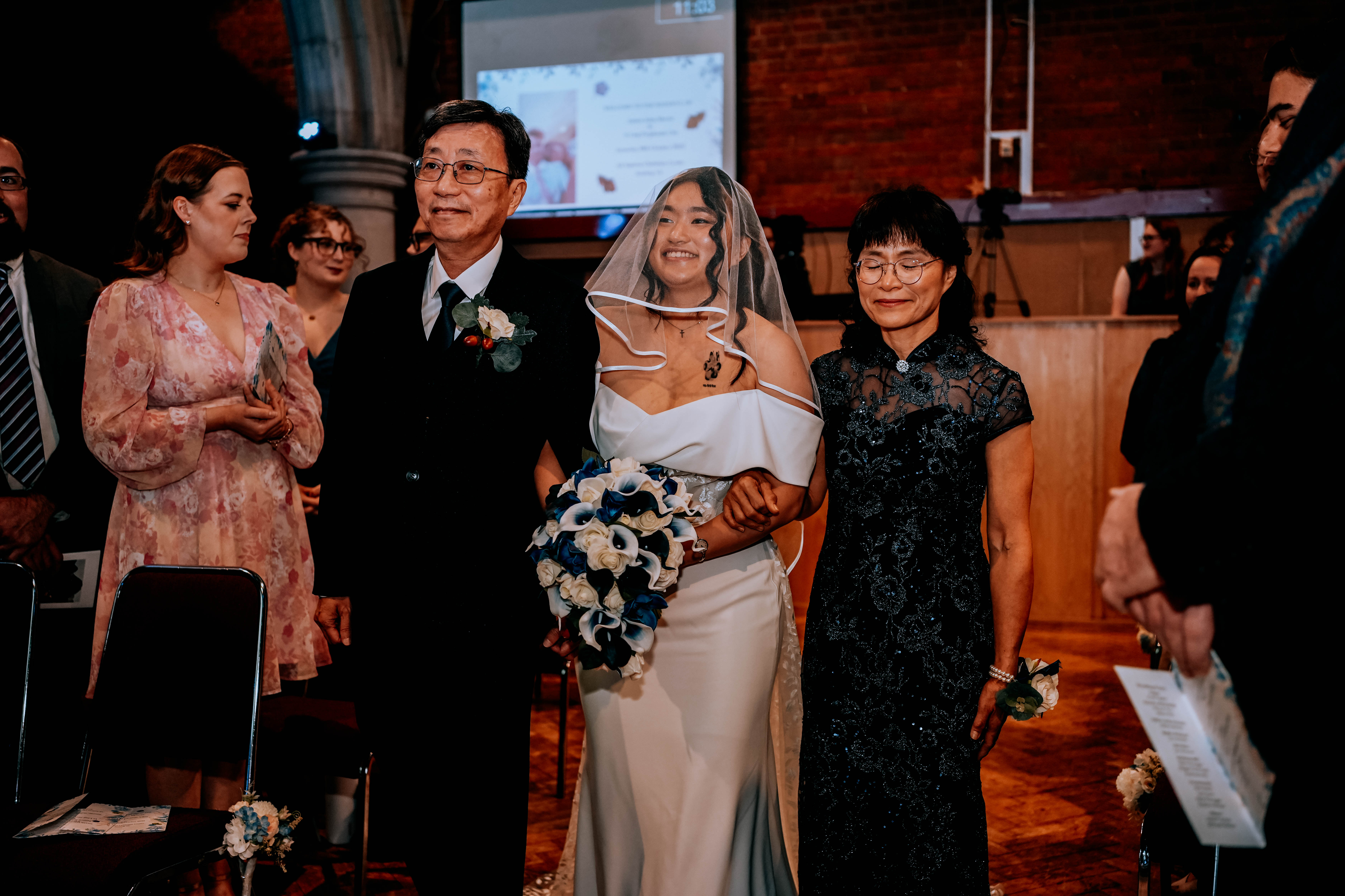 A smiling bride in a white off-the-shoulder gown and veil is escorted down the aisle by her parents. She holds her father's arm with one hand and a blue and white bouquet in the other, while her mother walks on her other side.