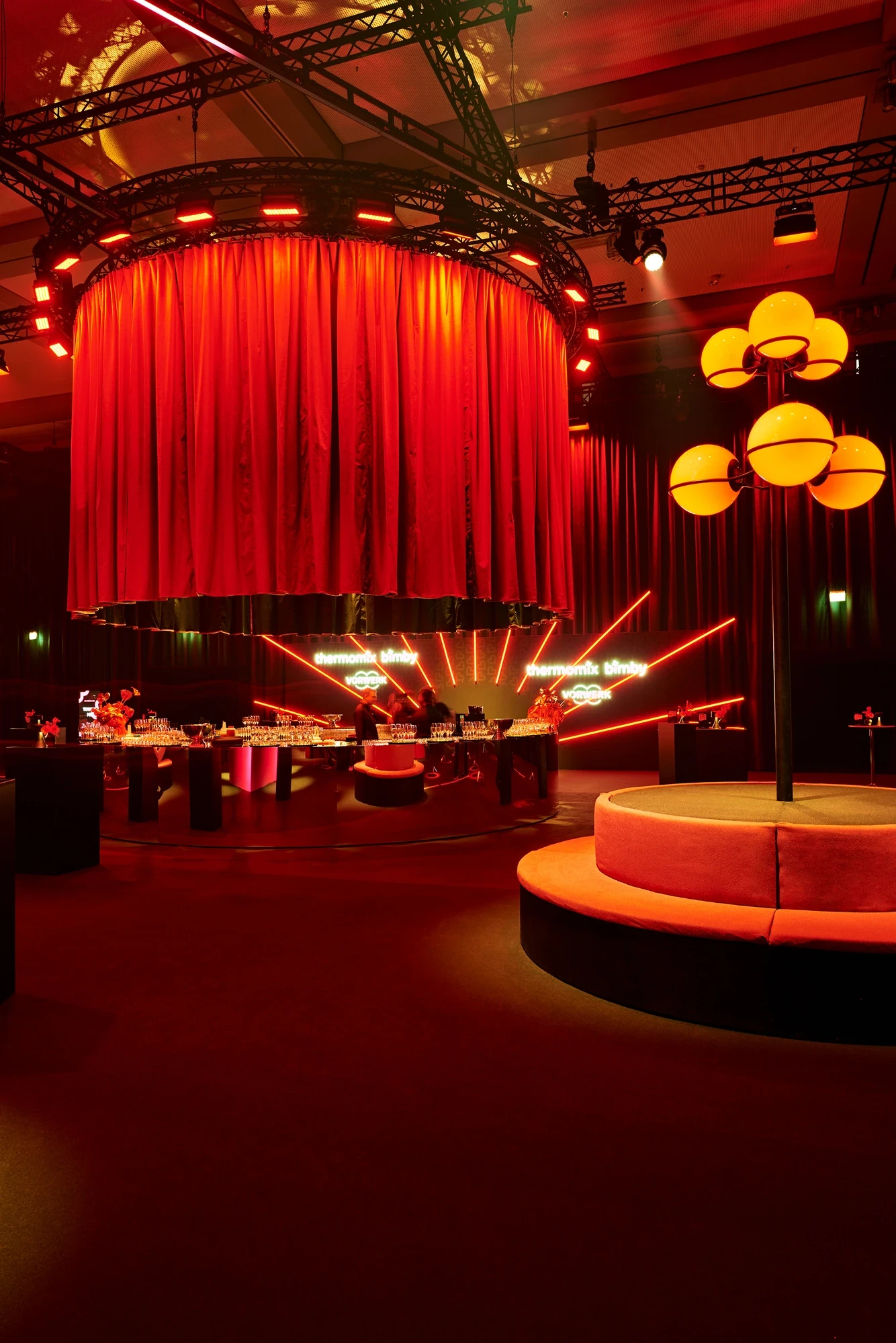 Vorwerk Lovebrand event with a circular red curtain canopy above the central stage, glowing Thermomix Bimby neon signs and a round seating arrangement in warm red light.