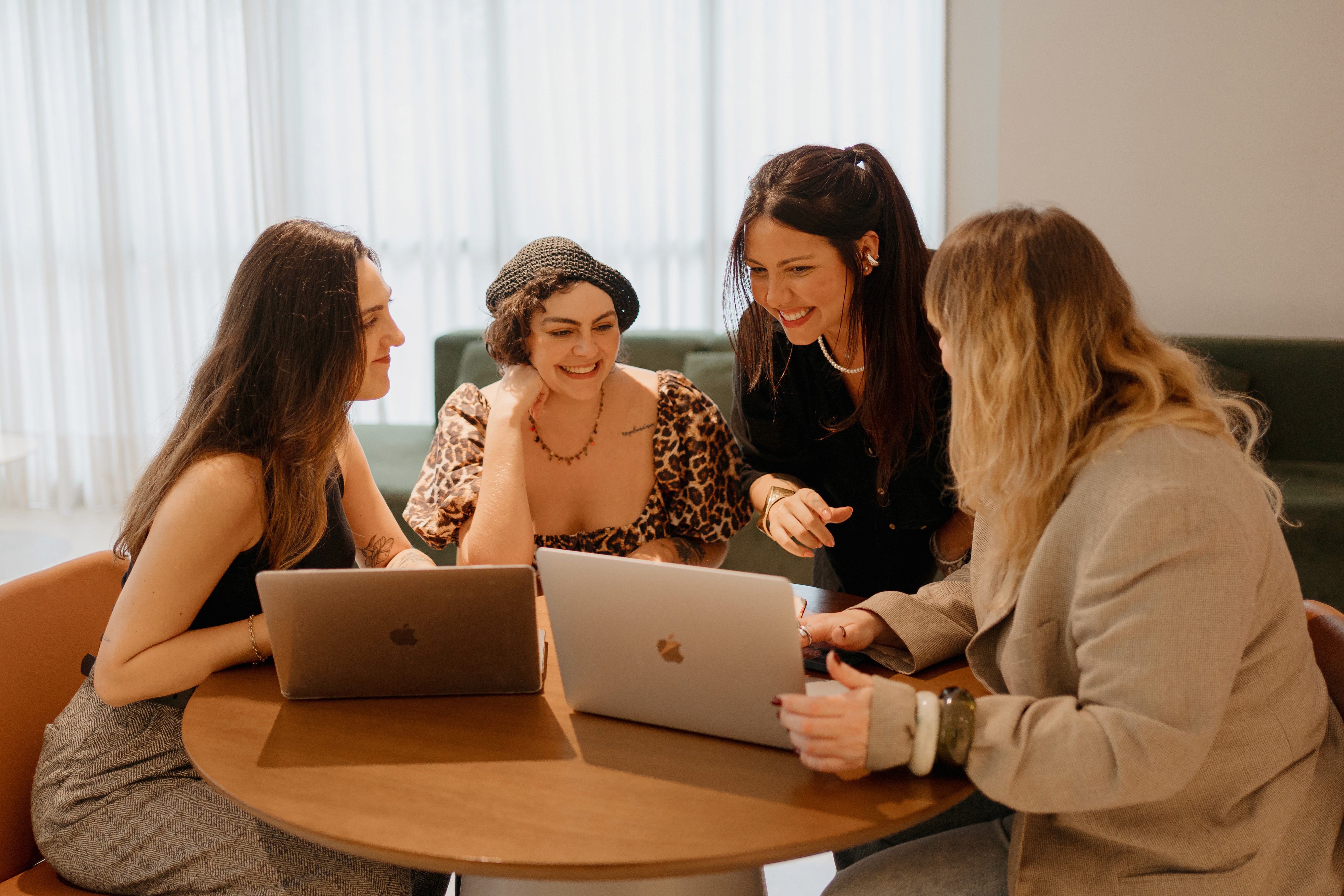 A young person with curly hair smiles at the camera while sitting in front of a laptop against a dark background.