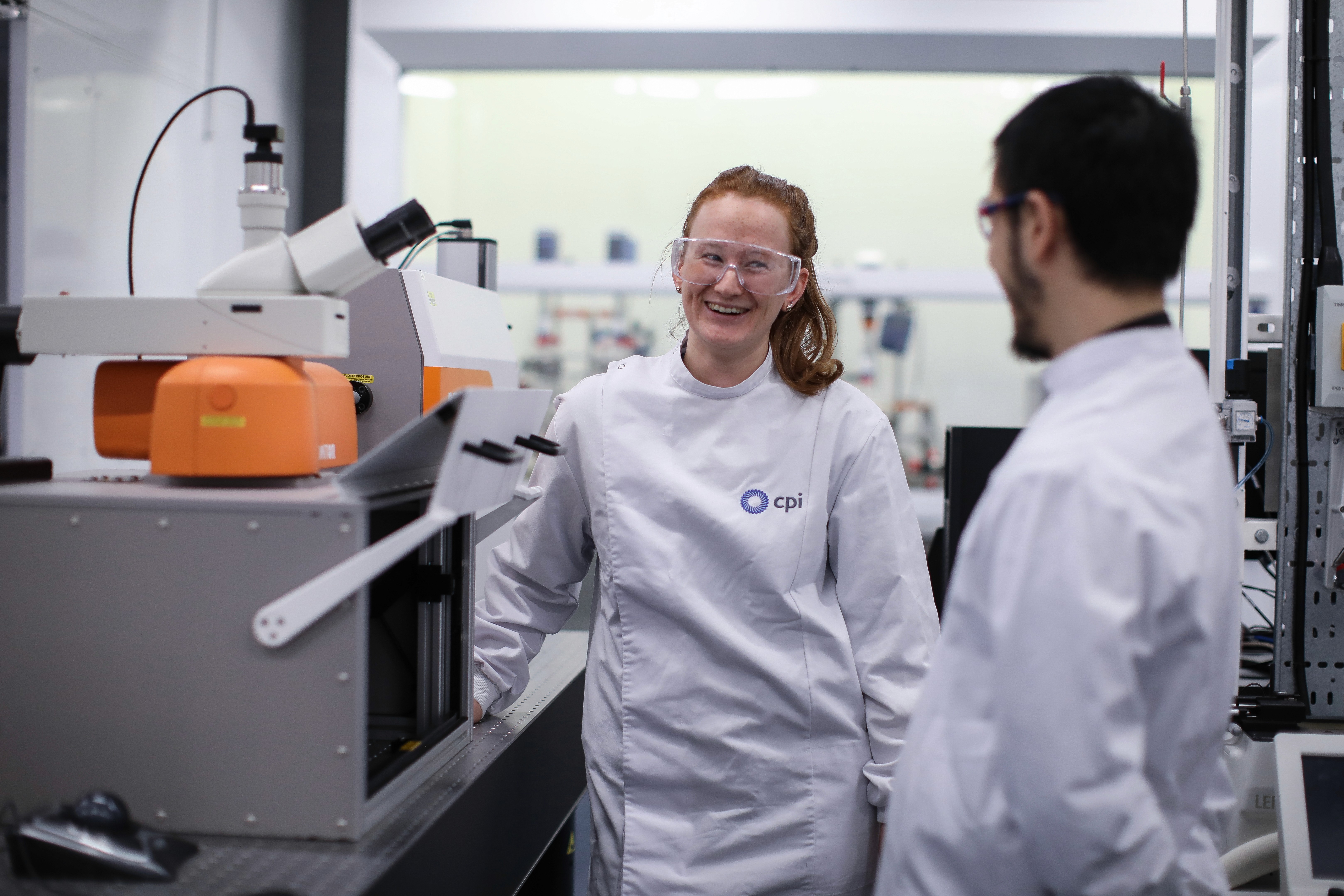 Smiling female scientist in a white CPI lab coat and safety glasses beside laboratory equipment, talking to a colleague