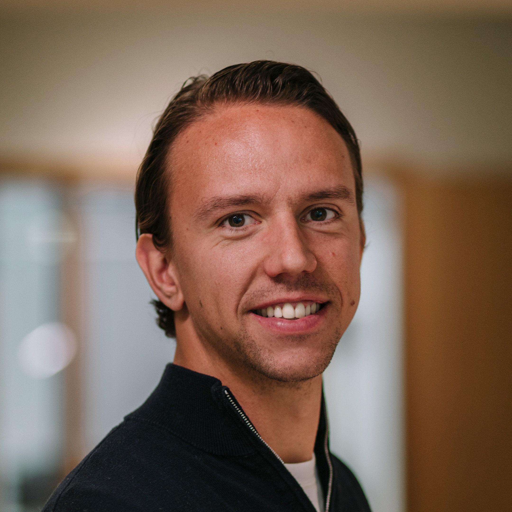 A smiling individual wearing a light shirt, posing for a photo in an indoor setting with blurred background.
