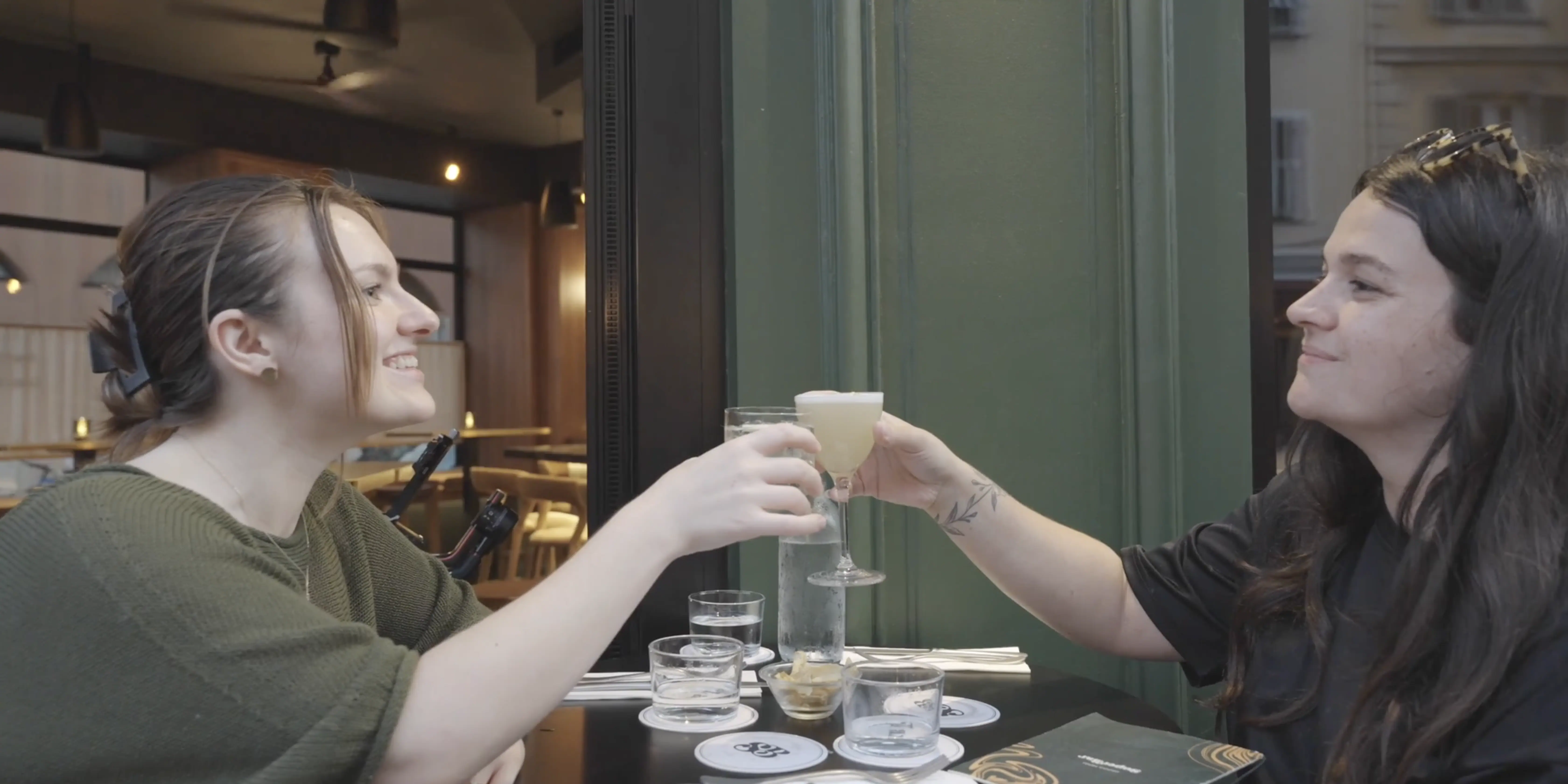 Two people clinking glasses at an outdoor café table, each enjoying a drink, with a cozy urban backdrop featuring blurred interior and exterior details.