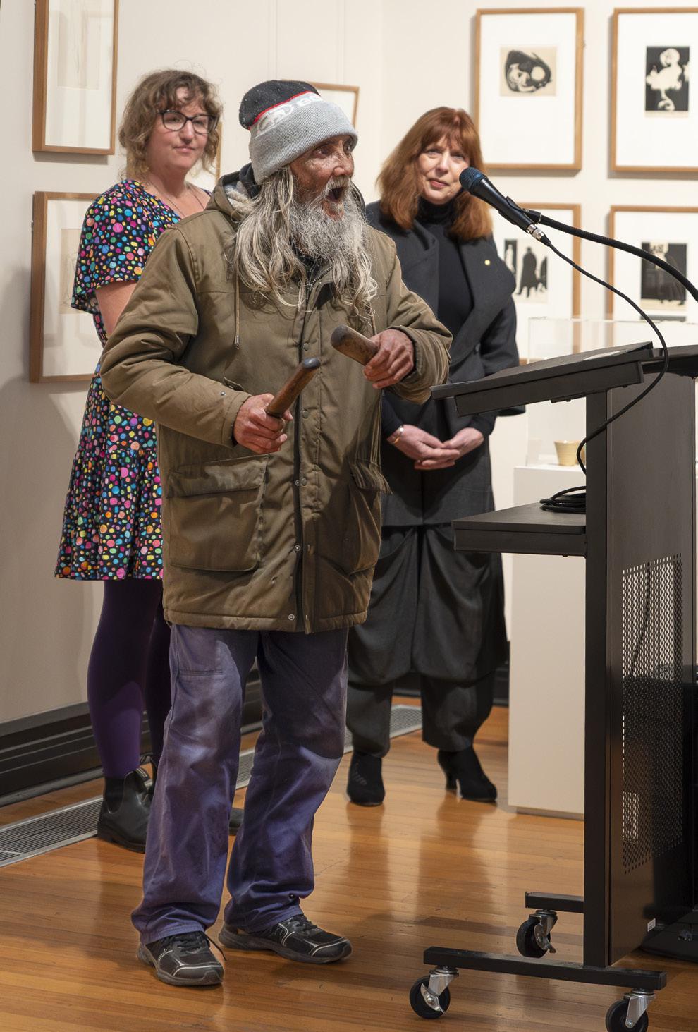 An elderly man in a winter coat and hat speaks into a microphone while holding an instrument, surrounded by two women in a gallery setting with framed artwork on the walls, showcasing a community art event.