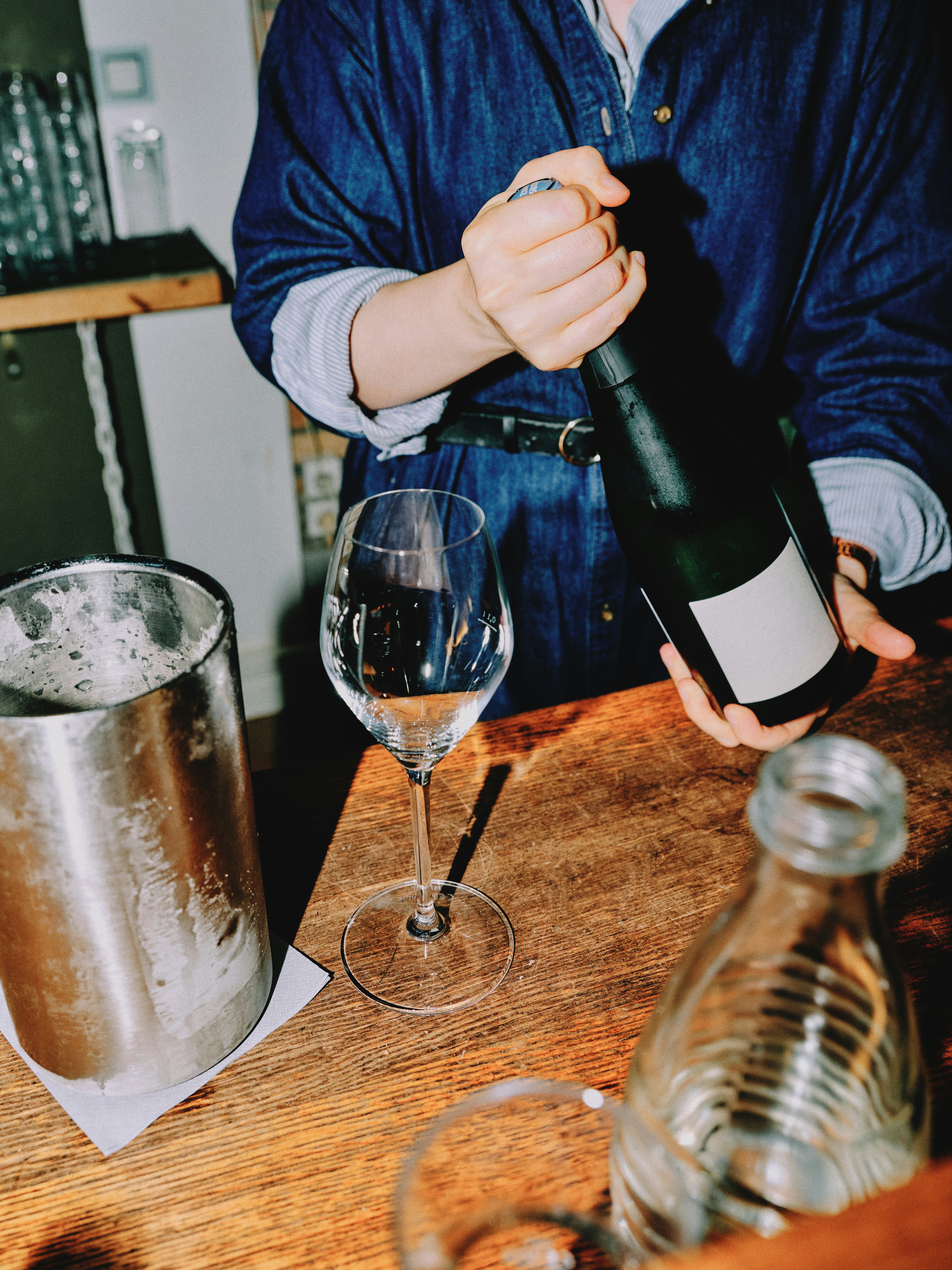 Close-up of a bartender in a denim dress holding a dark wine bottle beside a single stemmed wine glass on a wooden bar counter, silver ice bucket to the left