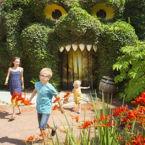 两个小孩和一个成年人走在一个巨大的、被植物覆盖的结构前,这个结构有眼睛和嘴巴。前景中有红花。
