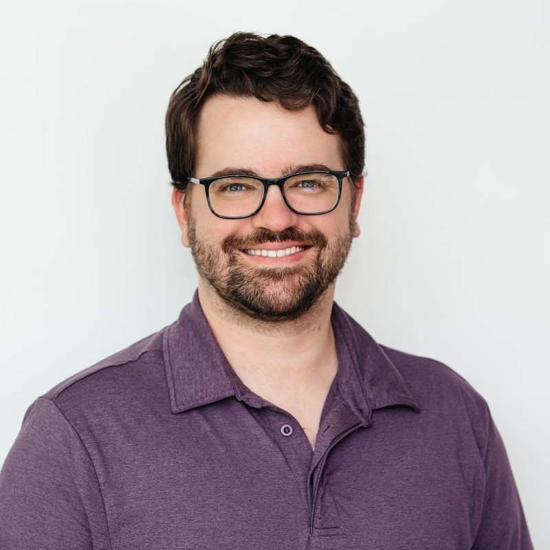 Close-up portrait of a freckled woman with soft pink hair and star earring, natural light, white tank top.