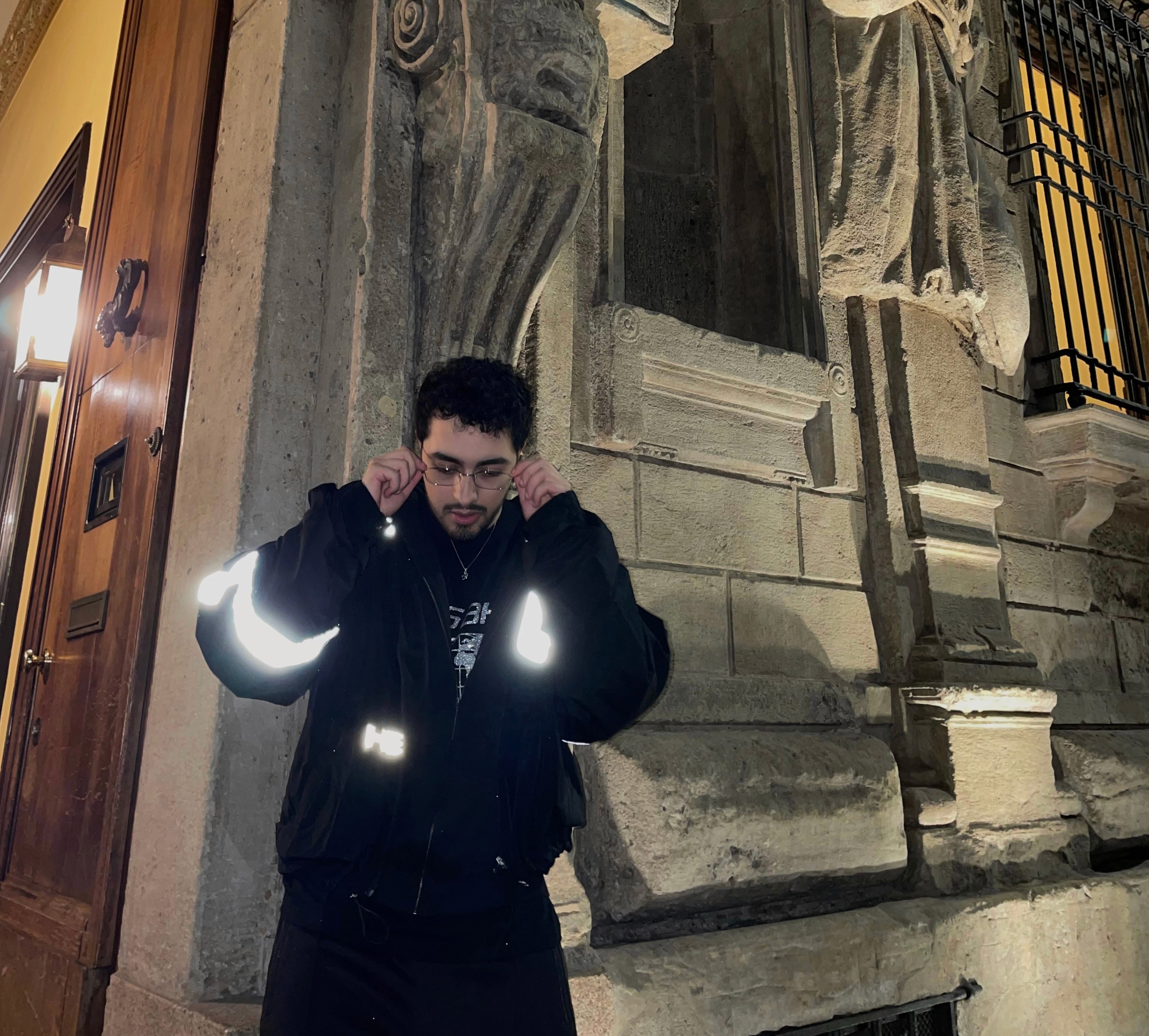 Italian producer Shadyboy stands outside a historic building at night, adjusting his glasses. He wears a black jacket with reflective patches, a hoodie, and a cross necklace, exuding a modern and stylish look.