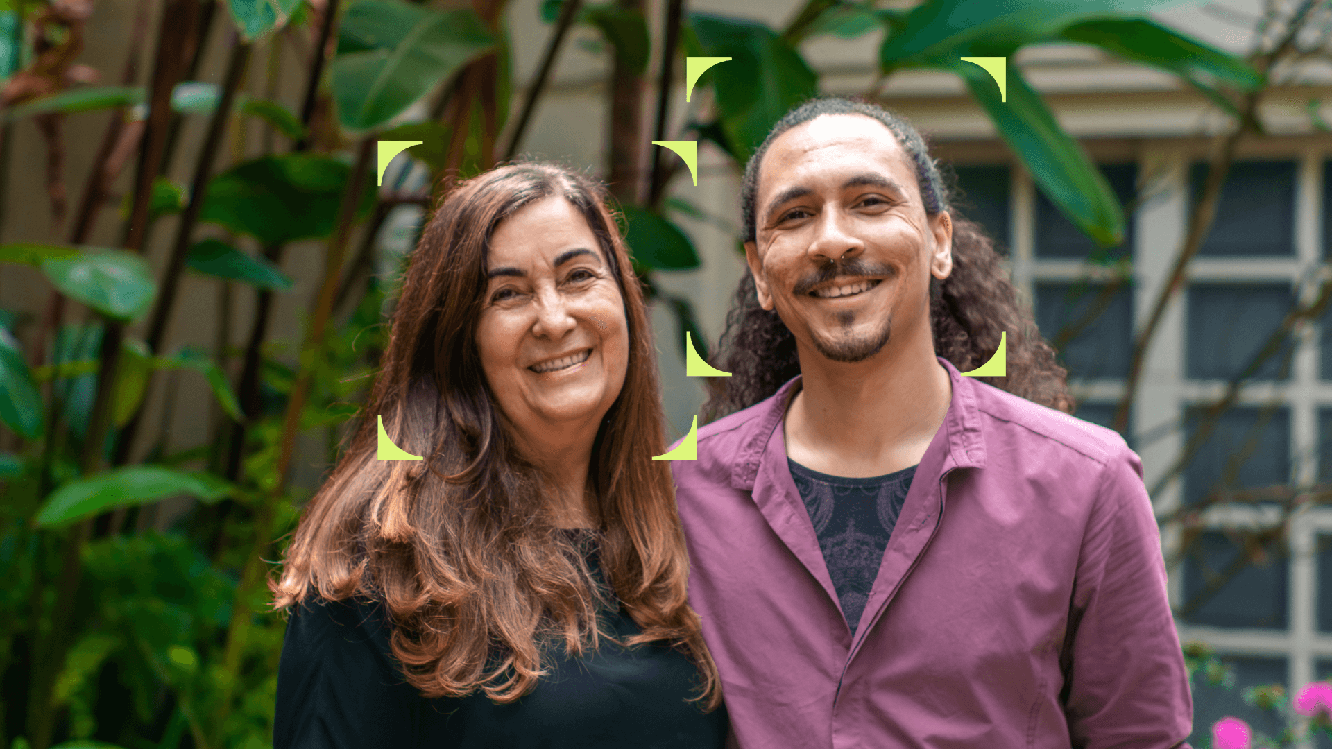 Foto de duas pessoas sorrindo lado a lado, em um ambiente externo com plantas ao fundo. À esquerda, uma mulher de cabelos longos e castanhos usa uma blusa preta; à direita, um homem de cabelos cacheados presos para trás usa uma camisa roxa. Ambos estão enquadrados por quatro marcadores gráficos em cantos arredondados, como se estivessem em destaque na imagem.