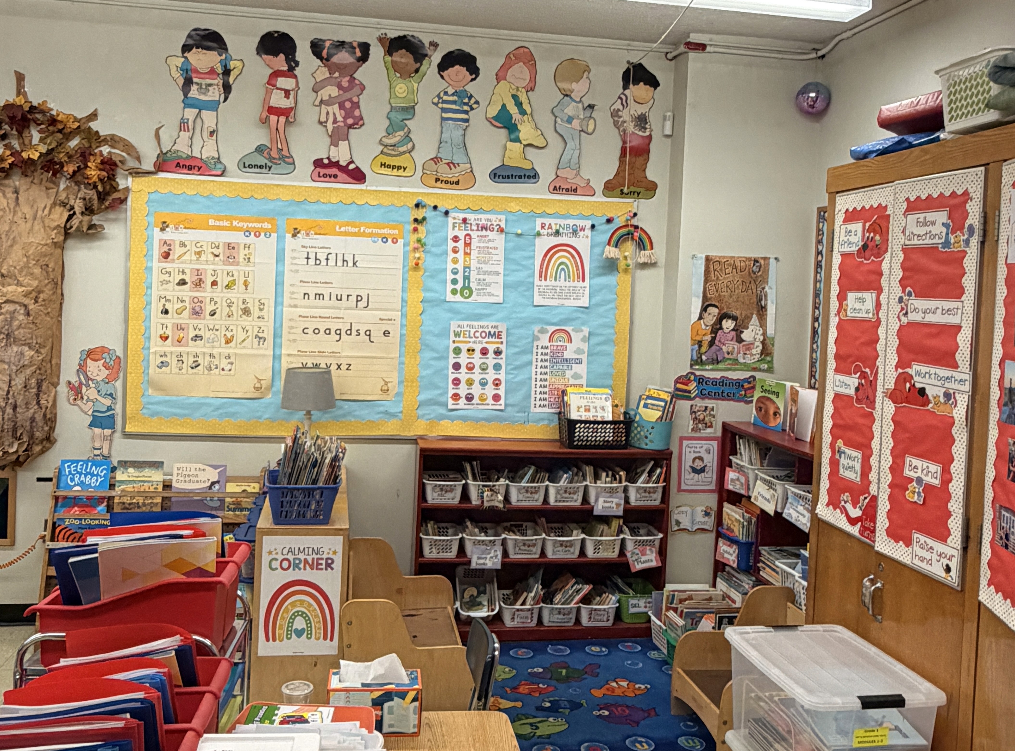 A classroom reading and calming corner with colorful bookshelves, bins of books, child-sized chairs, and a blue rug. The walls display alphabet and letter-formation posters, feelings charts, rainbow decorations, and illustrated emotion characters labeled “Angry,” “Lonely,” “Love,” “Happy,” “Proud,” “Frustrated,” “Afraid,” and “Sorry.” A cabinet on the right shows classroom rules such as “Be a friend,” “Follow directions,” “Do your best,” “Work together,” “Be kind,” and “Raise your hand.”