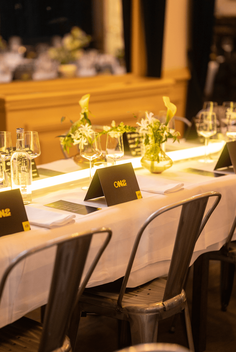 A smiling woman with her arms crossed, standing against a dark green background. She has long, dark hair.