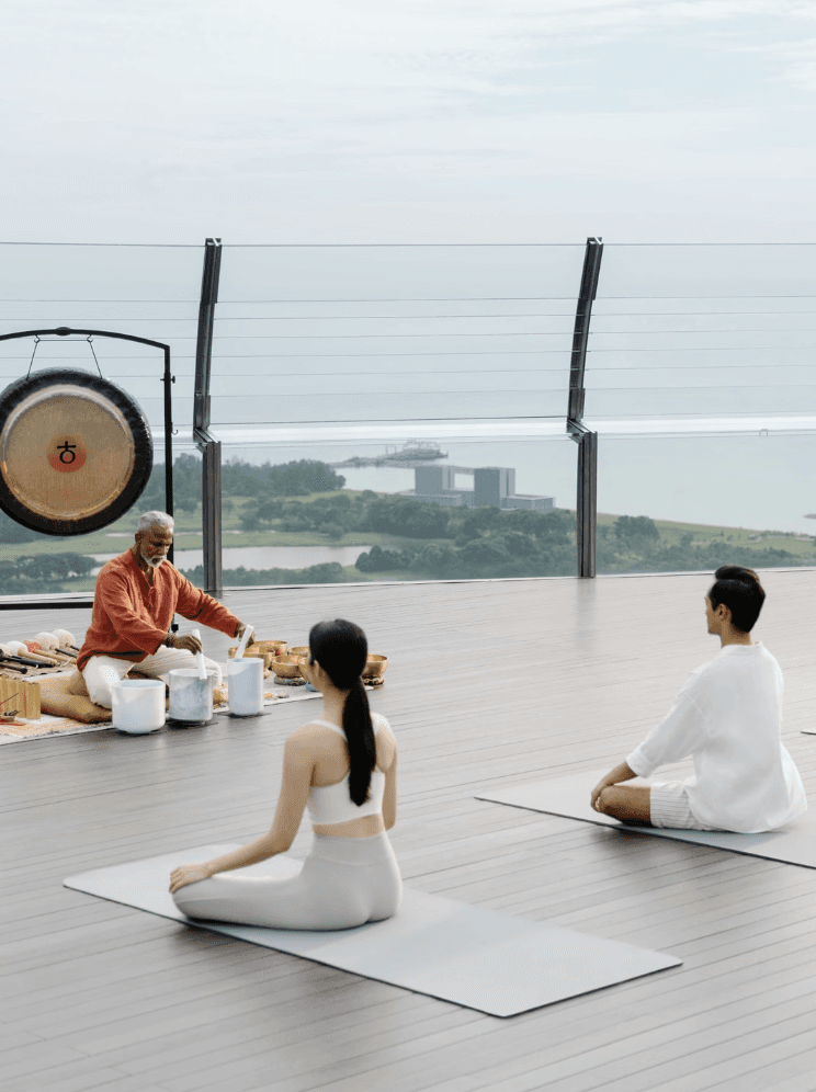 A serene yoga session taking place outdoors with a tranquil view. A teacher in traditional attire is leading two students seated on yoga mats, surrounded by various musical instruments, including a gong.
