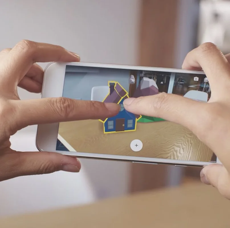 Hands holding smartphone with ARCore augmented reality mobile app displaying 3D house model overlay on table