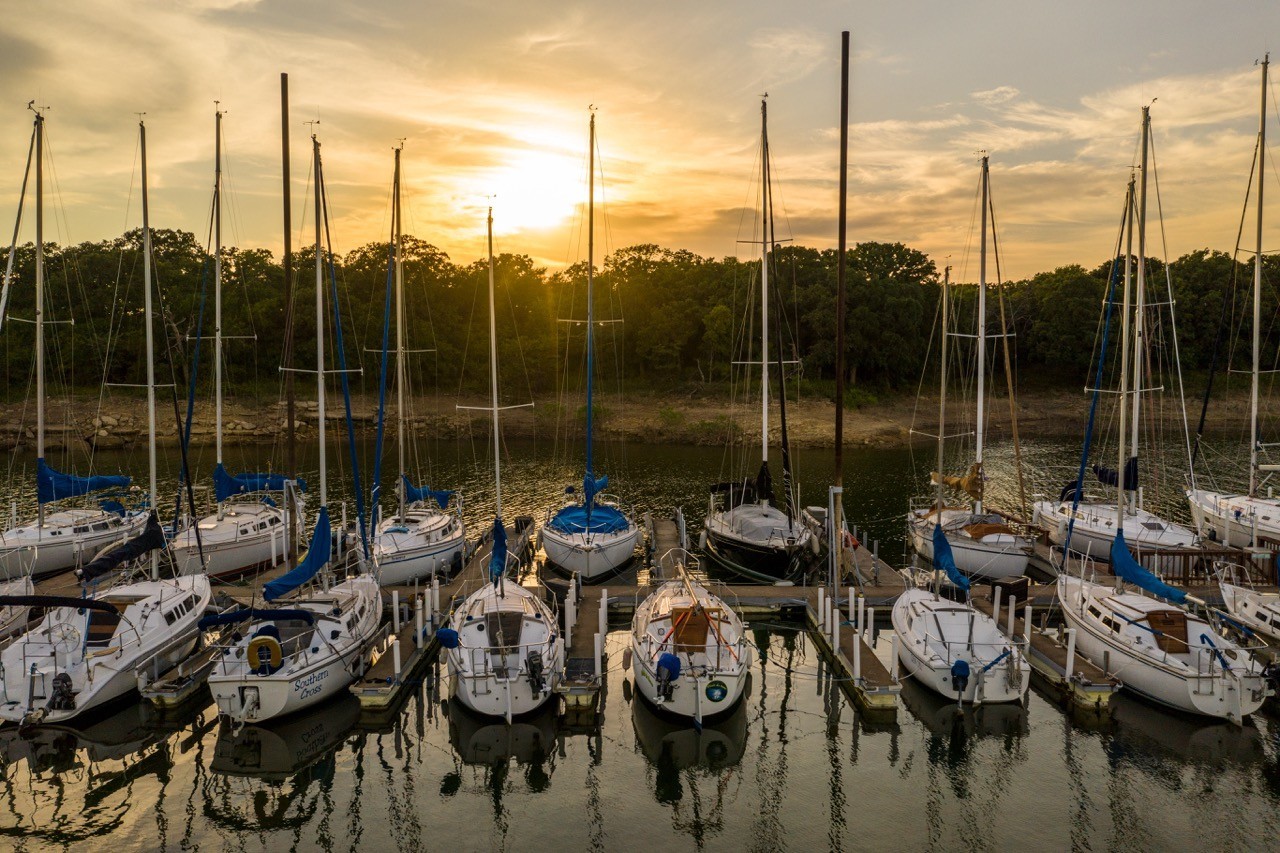 A tranquil marina at sunset with rows of docked sailboats and yachts reflecting on the calm water, surrounded by lush trees under a golden sky.
