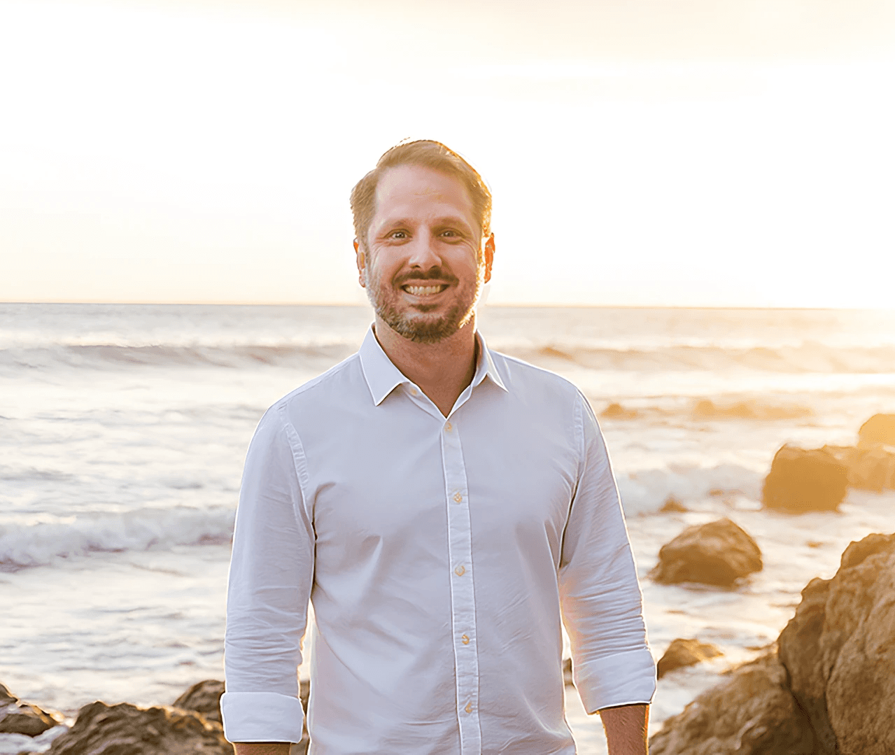 A smiling man stands on a beach at sunset, wearing a white shirt.