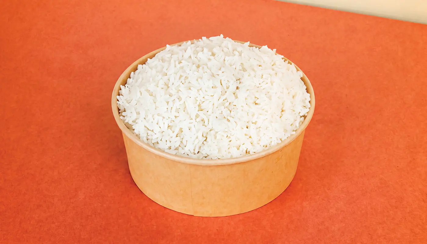 A round kraft-paper bowl filled to the top with fluffy white rice, photographed from above against a warm orange-red surface.