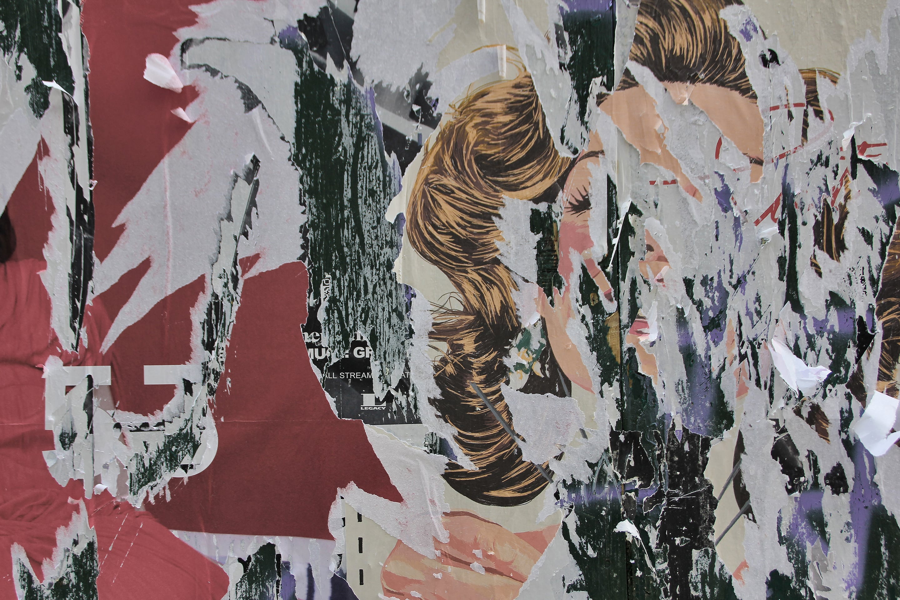 Photograph of a wall layered with torn posters revealing parts of a vintage-style illustration of a woman’s face and hair. The overlapping textures of red, white, and green paper create a fragmented, painterly surface evoking nostalgia and decay.