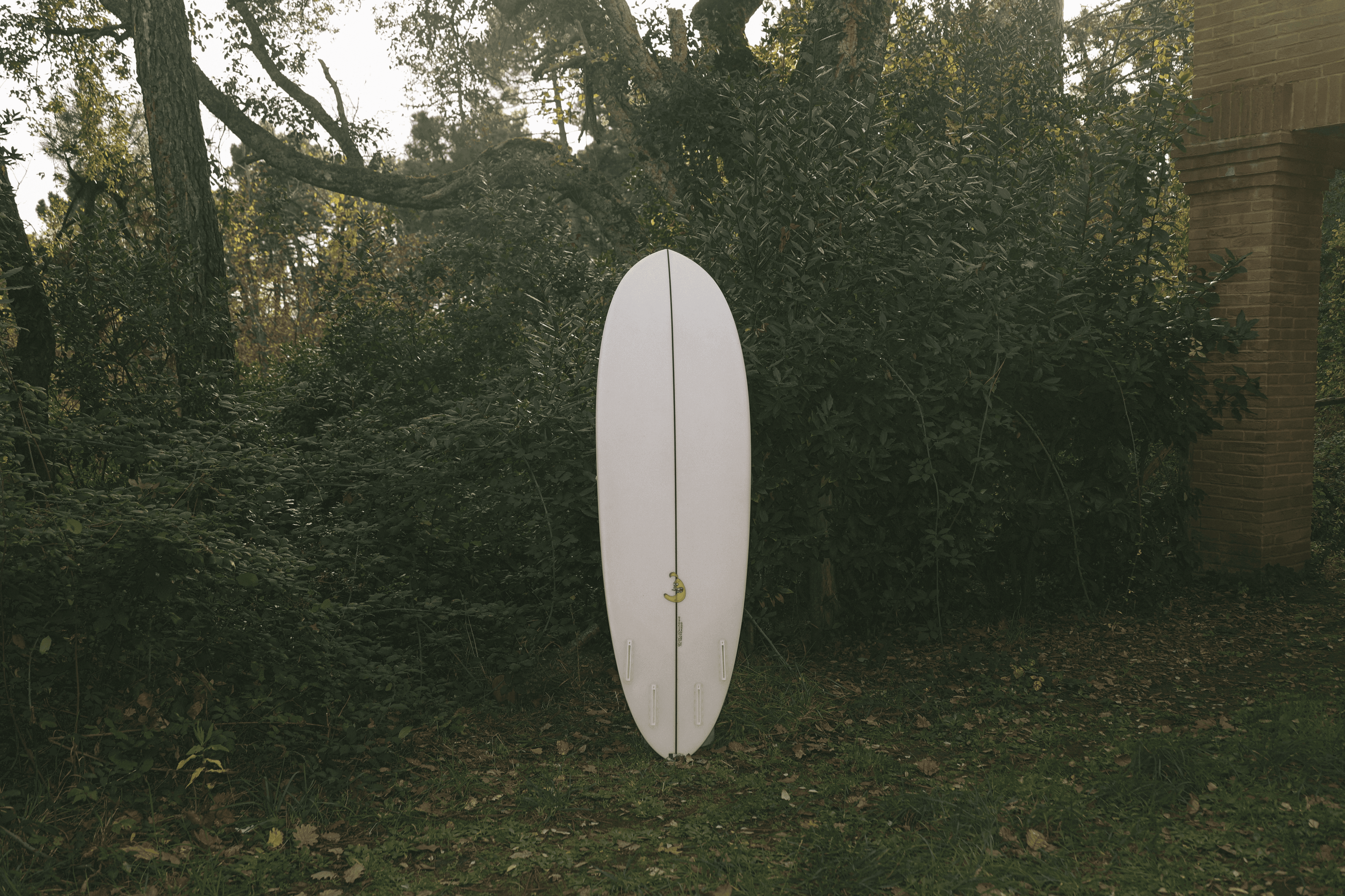 Tavola da surf modello Lakatan di colore bianco con pinna centrale gialla e configurazione thruster (tre pinne), esposta in piedi in un prato boschivo.