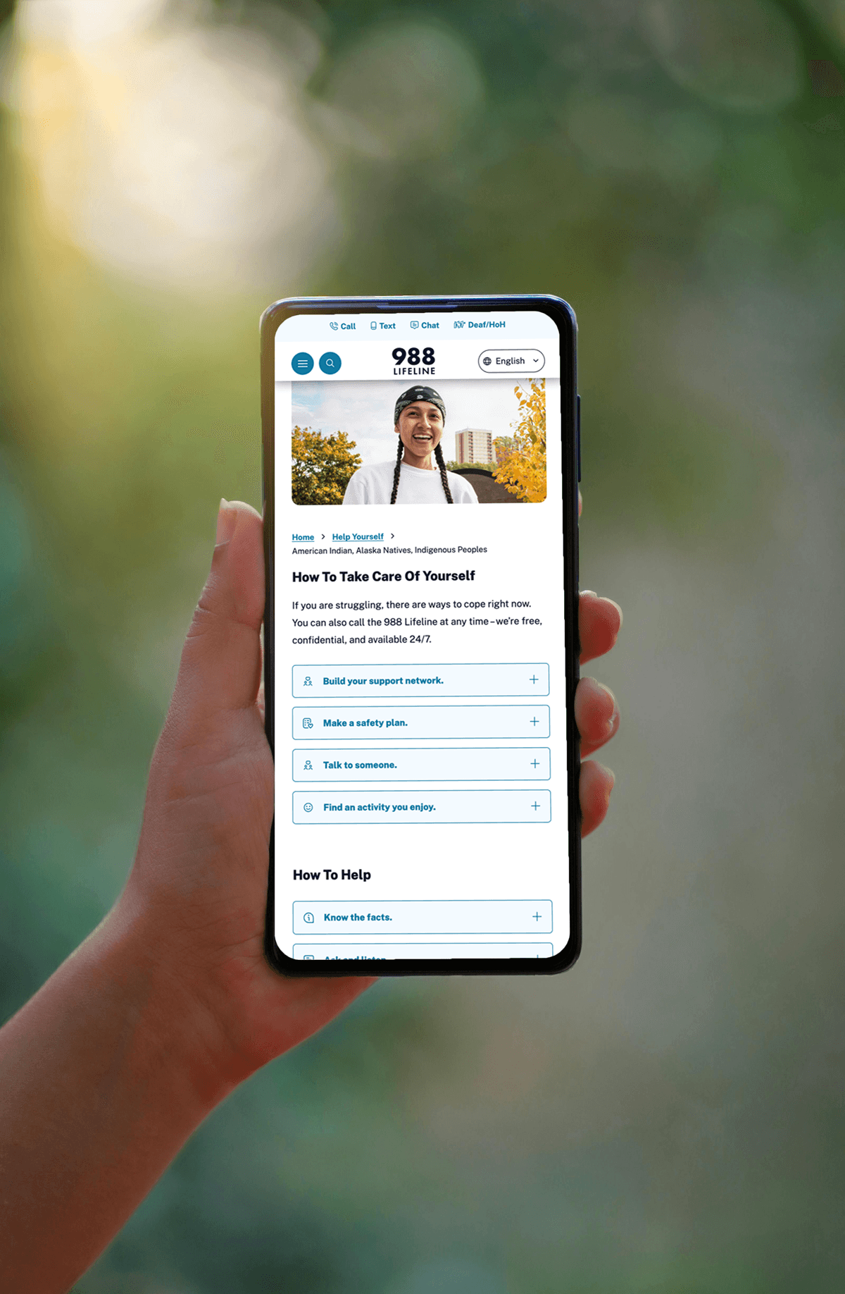 A hand is holding a mobile phone featuring a photo header and an accordion with tips on how to take care of yourself from the 988 website.