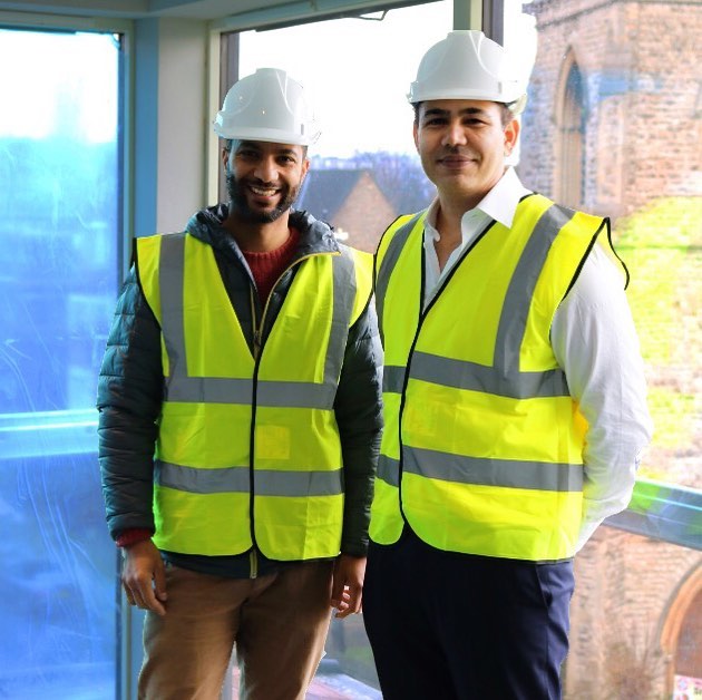Two professionals in a meeting, discussing ideas at a table with a laptop, notebook, and coffee cup in a modern office setting.
