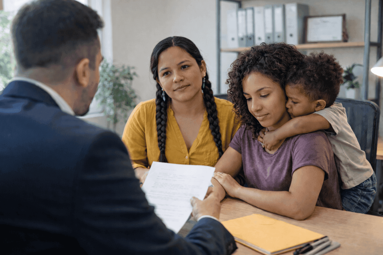 A young family consults with a professional in an office setting, as the individual in a suit discusses documents with them, while a child leans affectionately on one of the adults, conveying a sense of familial unity and serious discussion.