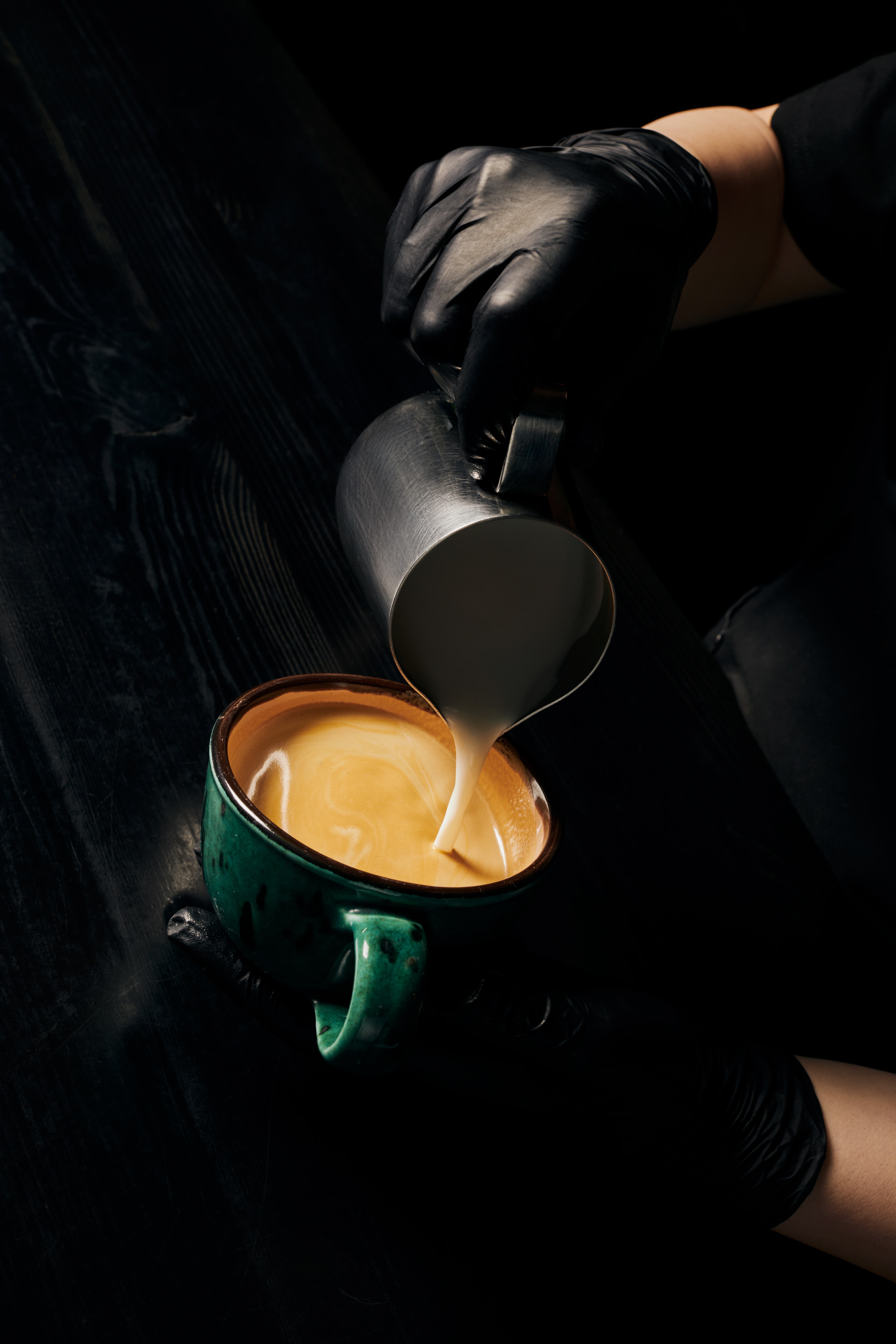 Close-up of a barista pouring steamed milk into a cup of coffee, captured in a dark, moody setting.