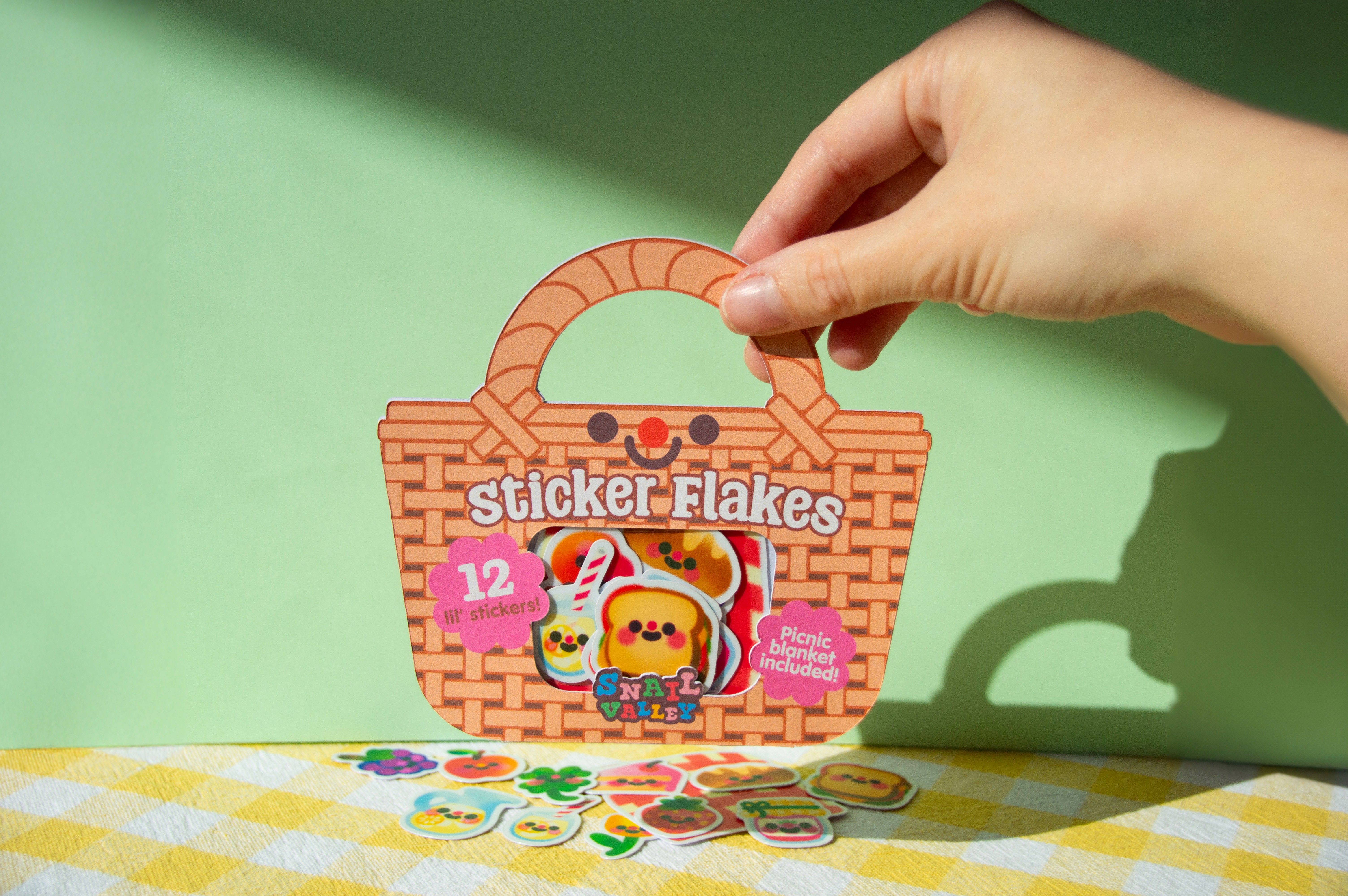 A hand holds a small package designed to look like a picnic basket. The basket has a smiling face and reads "Sticker flakes". There is a small window in the centre with tiny stickers of various picnic items are showing.