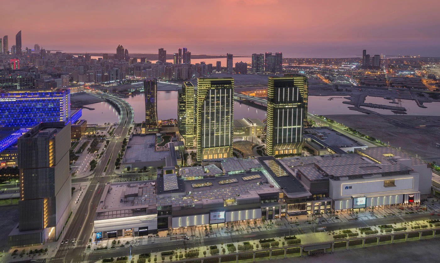 لقطة جوية نابضة بالحياة لجزيرة الريم أبوظبي وقت الغروب، تظهر الأبراج المضيئة ومركزاً تجارياً صاخباً مع انعكاس ألوان السماء الملونة على المياه المحيطة بالمنطقة السكنية الراقية