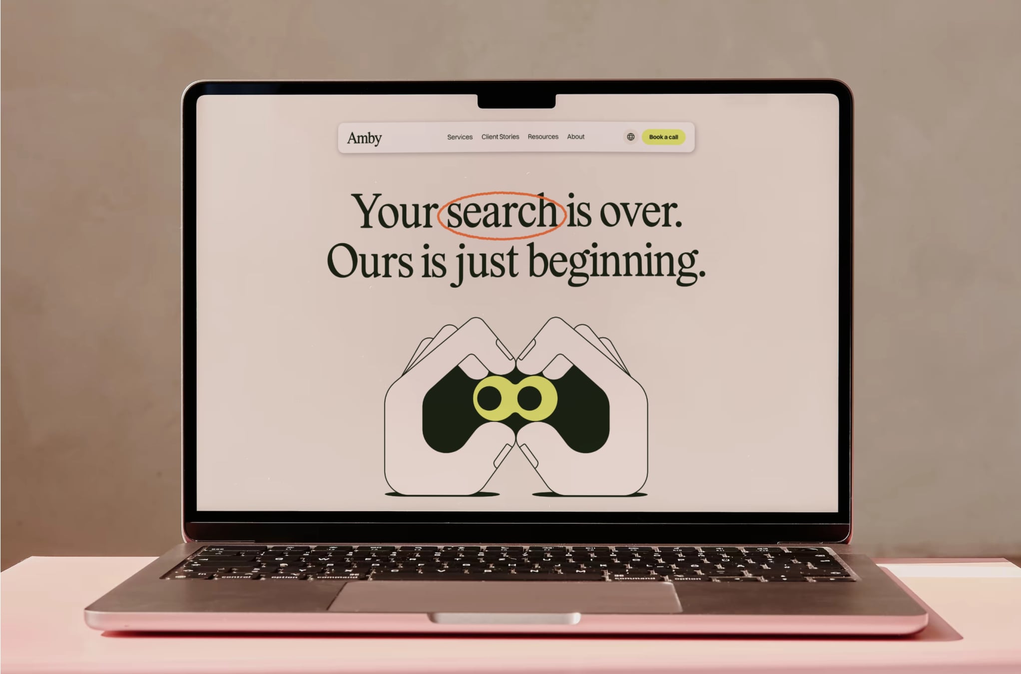 Laptop on a pink table showing the Amby website with the headline “Your search is over. Ours is just beginning.” above a binoculars illustration