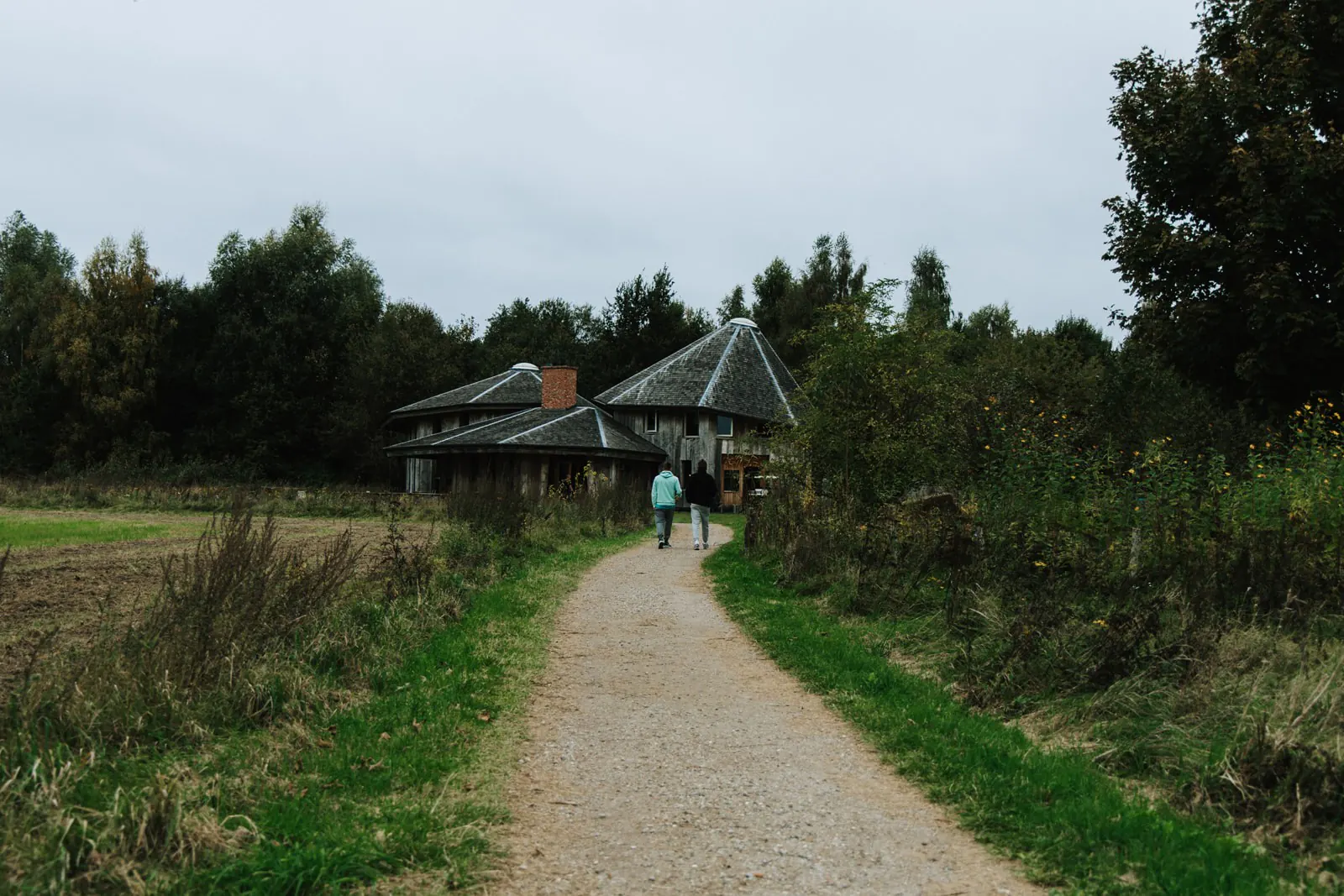 Twee wandelaars lopen over een grindpad tussen het groen naar een gebouw met puntdaken, onder een grijze lucht