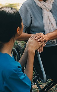  A female caregiver in blue scrubs sitting on the grass, holding the hands of a person sitting in a wheelchair