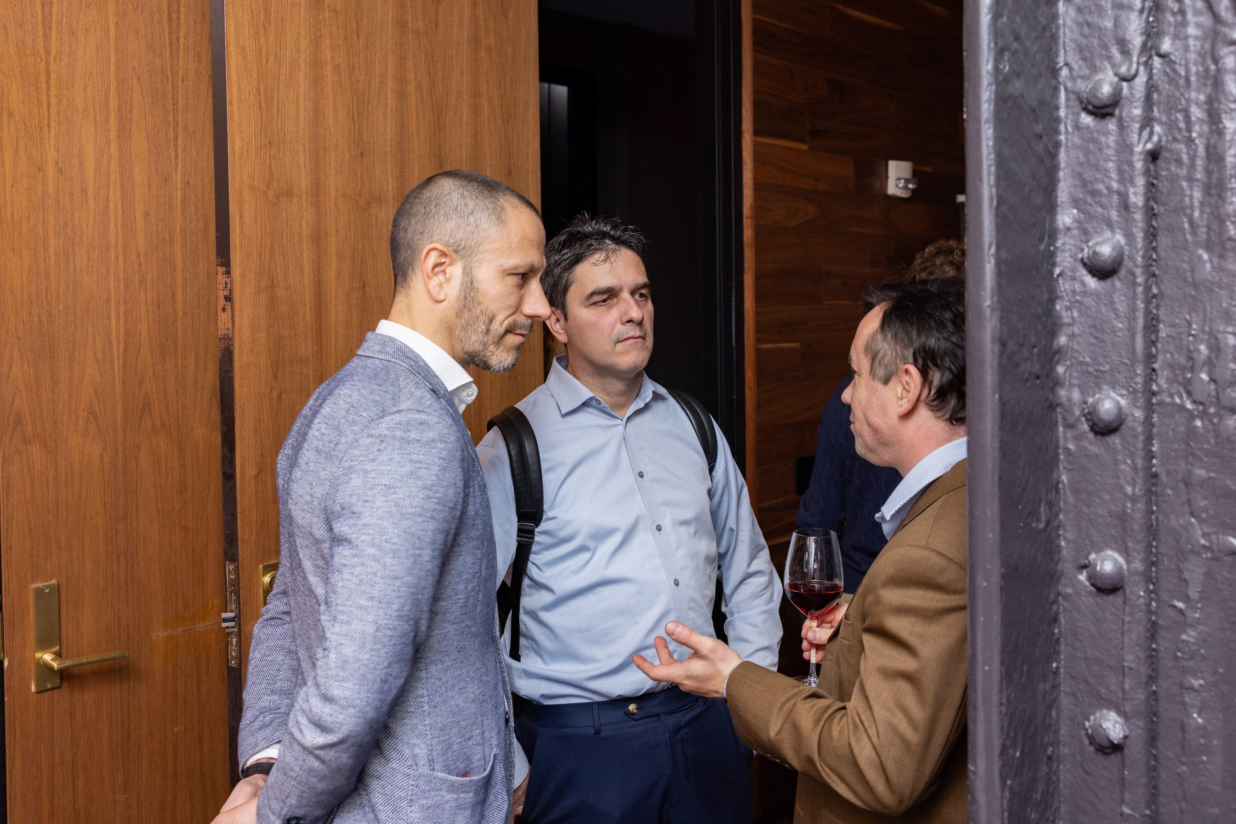 Three men engaged in a conversation near a doorway, with warm wooden tones in the background.