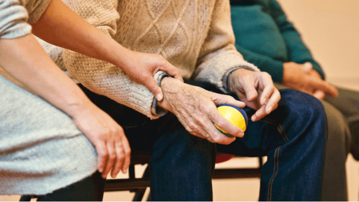 Caregiver supporting elderly person holding therapy ball.