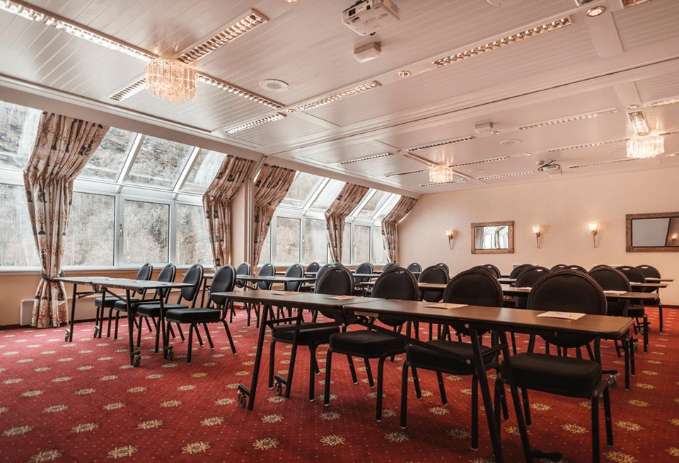 The image shows a spacious conference room with red carpet and tall windows, featuring rows of empty black chairs and tables under elegant ceiling lights.