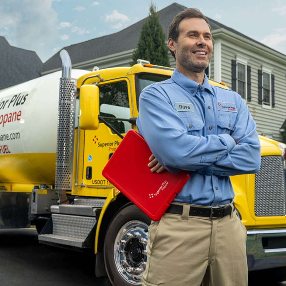 A man in a blue uniform stands in front of a yellow propane delivery truck, holding a red folder, outside a suburban house.