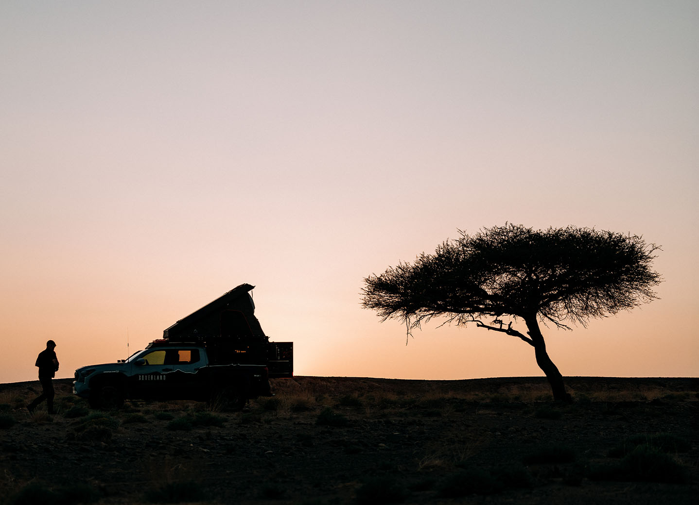 Silhouette of an overland vehicle with a rooftop tent and a person standing beside it on a ridge at sunset, with a lone tree to the right against a clear sky.